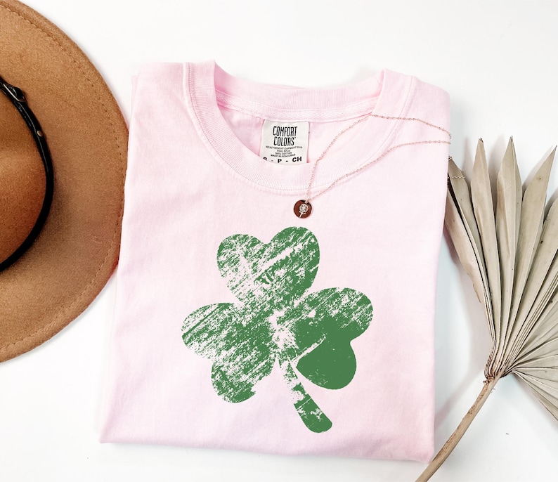 May include: Pink t-shirt with a green shamrock graphic. The shamrock has a distressed look and is centred on the shirt. The shirt has a crew neck and short sleeves.