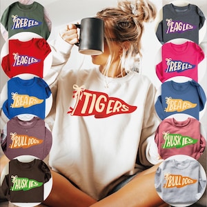 May include: A collection of crewneck sweatshirts and t-shirts in various colors, each featuring a team name on a pennant design. The sweatshirts and t-shirts are in colors like red, blue, green, and pink. The word "Tigers" is printed on a white sweatshirt.