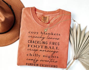 May include: A burnt orange short-sleeve t-shirt featuring black text that reads: cozy blankets, crunchy leaves, crackling fires, FOOTBALL, crisp mornings, chilly nights, comfy sweaters, PUMPKIN EVERYTHING. The shirt has a crew neckline.