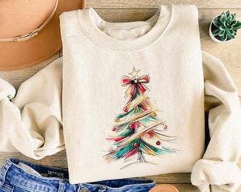 Op de afbeelding: Crèmekleurige sweatshirt met een kleurrijk kerstboomontwerp. De boom is versierd met rode, groene en gouden accenten, bekroond met een ster en een rode strik. De sweatshirt wordt gecombineerd met een blauwe spijkerbroek en een bruine hoed.