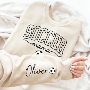 Könnte beinhalten: Beiges Sweatshirt mit dem Text "SOCCER mama" in schwarzen Blockbuchstaben. Der Ärmel zeigt den Namen "Oliver" in Schreibschrift mit einer Fußballgrafik. Das Sweatshirt ist aus weichem Material.