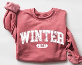 May include: A red-pink sweatshirt with the word "WINTER" in large, distressed white letters across the front. Below the word "WINTER" is the word "VIBES" in a smaller, white, oval-shaped font. The sleeves are rolled up.