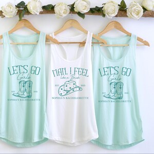 Puede incluir: Tres camisetas sin mangas con un tema de despedida de soltera. La camiseta blanca dice "Man, I feel like a bride" con un gr&aacute;fico de sombrero de vaquero. Las dos camisetas sin mangas azul claro dicen "Let's go girls" con un gr&aacute;fico de un par de botas de vaquero. Las tres camisetas sin mangas tienen el texto "Sophia's Bachelorette" impreso en ellas.