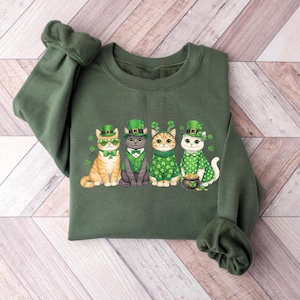 May include: Green sweatshirt featuring four cartoon cats dressed for St. Patrick's Day. Each cat wears a green hat and is adorned with shamrocks. One cat has green heart-shaped glasses, and another has a pot of gold.