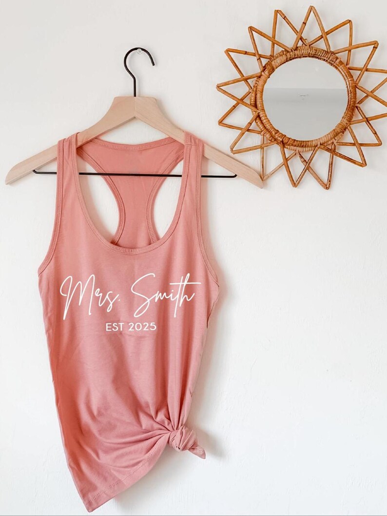 Pu&ograve; includere: Un top rosa polvere con una grafica bianca che dice "Mrs. Smith EST 2025" appeso a un appendiabiti di legno.