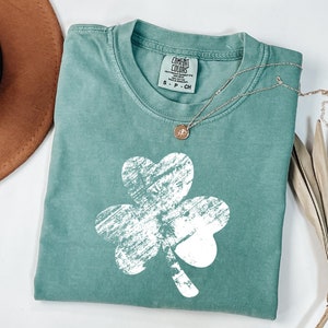 May include: A teal green t-shirt featuring a white distressed shamrock design. The shirt has a crew neck and is made from a soft, comfortable fabric.