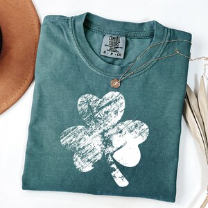 May include: A teal green t-shirt with a white distressed shamrock design. The shirt has a crew neck and short sleeves. The brand label reads "Comfort Colors" with the size "S-P-CH".
