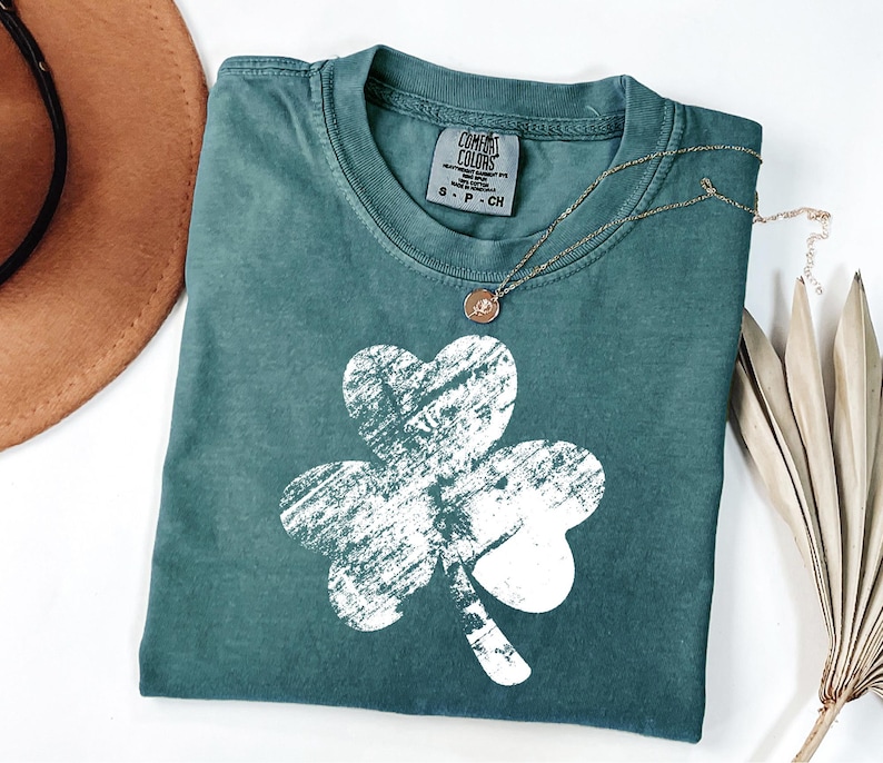 May include: A teal green t-shirt with a white distressed shamrock design. The shirt has a crew neck and short sleeves. The brand label reads "Comfort Colors" with the size "S-P-CH".