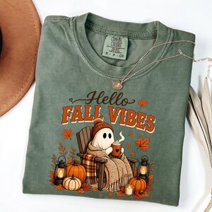 May include: A sage green t-shirt featuring a graphic of a ghost wearing a beanie and wrapped in a blanket, sitting in a wooden chair with a mug. The text reads Hello FALL VIBES, surrounded by pumpkins, lanterns, and autumn leaves in orange and brown.