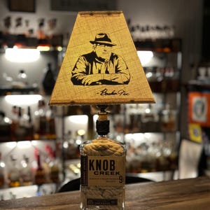 May include: A unique lamp made from a Knob Creek whiskey bottle. The lampshade features a black and white illustration of a man in a hat. The bottle is wrapped with rope and has a label that reads "Knob Creek 9".