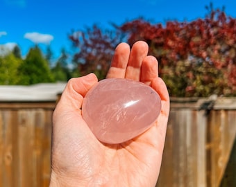 Può includere: Un grande cristallo di quarzo rosa a forma di cuore, liscio e traslucido, tenuto in una mano. Il cristallo è rosa pallido e traslucido. Lo sfondo è un'immagine sfocata di una recinzione di legno e alberi.