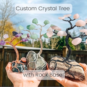 Könnte beinhalten: Drei individuell gestaltete Kristallbäume mit Steinsockeln. Die Bäume sind aus Kupferdraht und verschiedenen Kristallen gefertigt, darunter Amethyst, Rosenquarz und grüner Aventurin. Die Steinsockel sind natürlich und haben eine graue und braune Farbe.