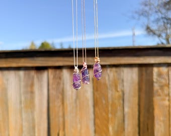 May include: Three amethyst crystal necklaces hanging in front of a wooden fence. Each pendant is a raw, purple crystal wrapped in wire and suspended from a delicate chain. The background features a clear blue sky and a blurred view of trees.