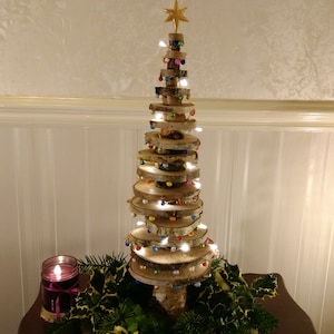 Może przedstawiać: Choinka drewniana wykonana z ułożonych na sobie okrągłych kawałków drewna brzozowego, ozdobiona kolorowymi światełkami i koralikami. Choinka stoi na stole z świecą i zielenią.