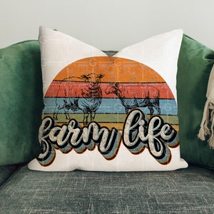 Puede incluir: Almohada blanca con un gr&aacute;fico de estilo vintage de dos ovejas frente a una puesta de sol con el texto "farm life" en una fuente retro.
