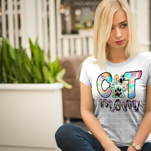 以下が含まれることがあります： 「Cat Mom」と書いてあるカラフルなタイダイ柄の白いTシャツ。Cの真ん中に肉球のプリントがあります。