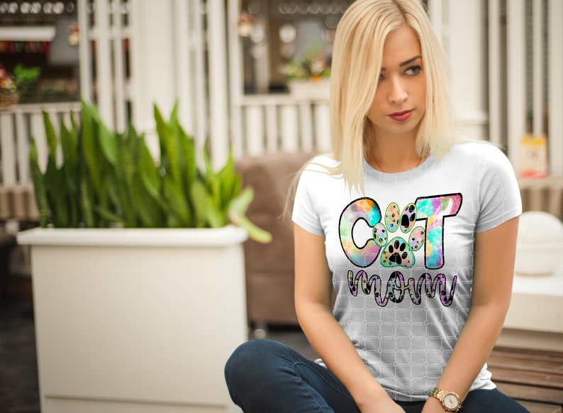 以下が含まれることがあります： 「Cat Mom」と書いてあるカラフルなタイダイ柄の白いTシャツ。Cの真ん中に肉球のプリントがあります。