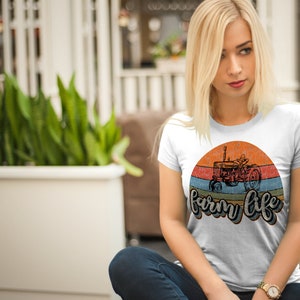 May include: White t-shirt with a vintage-style graphic of a tractor in front of a sunset with the text "farm life" in a retro font.
