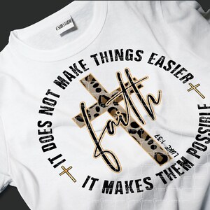 May include: White t-shirt with a black and gold graphic design. The graphic features a leopard print cross with the word "faith" written in a gold script. The text around the cross reads "It does not make things easier + It makes them possible" with the Bible verse "Luke 1:37" underneath.