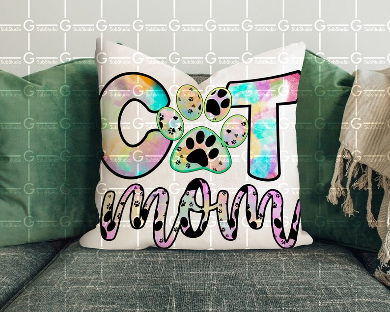 以下が含まれることがあります： 猫の足跡がついた「Cat Mom」と書かれたカラフルなタイダイ柄の白い枕。