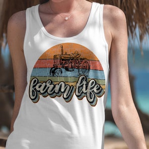 May include: White tank top with a vintage-style graphic of a red tractor in front of a sunset with the text "farm life" in a retro font.
