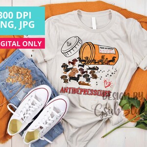 May include: A white t-shirt with a graphic of a pill bottle spilling out dachshund dogs. The text on the pill bottle reads "Pilules du bonheur" and the text below the graphic reads "Antidepresseurs".