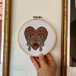 Puede incluir: Un corazón marrón con una cara triste y ojos en forma de lágrima está bordado en tela blanca en un aro de madera circular.