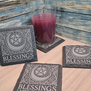 May include: A set of four square slate coasters with a crescent moon and pentagram design, surrounded by ornate scrollwork. The word "BLESSINGS" is etched below the design in a serif font. A tall glass with a purple liquid sits on one of the coasters.