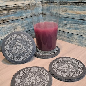 May include: A set of four round slate coasters with a Celtic knot design. The coasters are dark gray with a white, intricate knot pattern. A glass of purple liquid sits on one coaster. The background is a blue and brown wood grain.