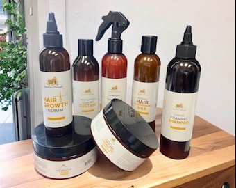 May include: A set of hair care products in brown glass bottles with black lids and white labels. The products include hair growth serum, curling custard, hair milk, and liquid foaming shampoo. The labels feature the brand name "Nature's Essentials" and product descriptions. There is also a jar of hair oil.