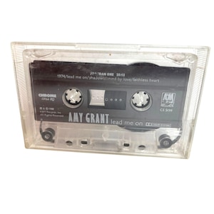 May include: A clear plastic cassette tape case containing a black audio cassette. The label features the text Amy Grant Lead Me On, A&M Records, and track listings including 1974, Lead Me On, Shadows, Saved By Love, and Faithless Heart.