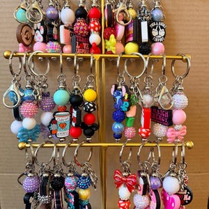 May include: A display of beaded keychains with various charms. Keychains feature colorful beads, bows, and charms with text like "Teach Love Inspire" and "Match Energy". The keychains are displayed on a gold-colored metal rack.