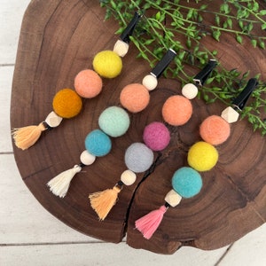 Op de afbeelding: Vijf kleurrijke viltballen haarclips met houten kralen en kwastjes. De kleuren zijn geel, oranje, perzik, lichtgroen, turquoise, grijs, roze en lichtblauw.