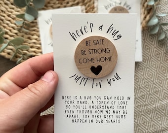 May include: A white card with black text that reads "here's a hug just for you" with a wooden circle attached to the card. The circle has the text "Be Safe Be Strong Come Home" and a small black heart.