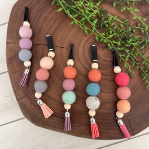 May include: Five colorful car air fresheners with felt ball accents and tassels. Each freshener features a black clip, wooden beads, and a series of felt balls in various pastel colors, including pink, purple, orange, and blue. The tassels are in coordinating colors.