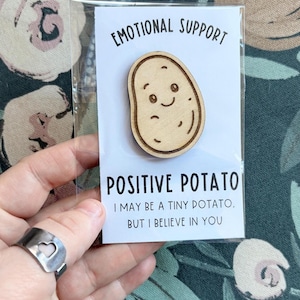May include: A wooden pin shaped like a potato with a smiling face, attached to a white card in a clear plastic sleeve. The card features the text: EMOTIONAL SUPPORT, POSITIVE POTATO, I MAY BE A TINY POTATO, BUT I BELIEVE IN YOU, printed in black ink.