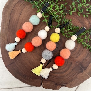 Op de afbeelding: Drie kleurrijke viltballenkettingen met houten kralen en kwastjes. Elke ketting heeft een zwarte clip om te bevestigen aan een kinderwagen, luiertas of andere items.