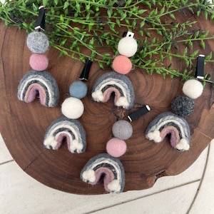 Op de afbeelding: Zes regenboog haarclips van vilt met viltballen in verschillende kleuren, waaronder grijs, wit, roze en blauw. De clips zijn bevestigd aan de viltballen met zwarte clips.