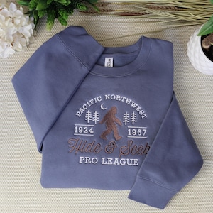 Peut inclure: Un sweat-shirt bleu ardoise à col rond avec une broderie représentant une silhouette de Bigfoot marron, des sapins et un croissant de lune. Le texte indique Pacific Northwest, 1924, 1967, Hide & Seek et Pro League en fil blanc et marron.
