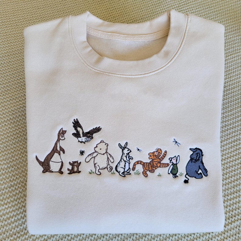 Op de afbeelding: Cr&egrave;mekleurige sweatshirt met geborduurde figuren uit een kinderboek. Het ontwerp bevat een kangoeroe, een beer, een konijn, een tijger, een varkentje en een ezel. De figuren zijn in verschillende kleuren, met een eenvoudige, grillige stijl.