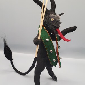 Può includere: Una figura artigianale di Krampus in feltro nero con corna, una lunga coda e una lingua rossa. Indossa una sciarpa verde con campanelle d'argento e tiene due bastoncini di legno. La figura ha un viso grigio e occhi gialli.