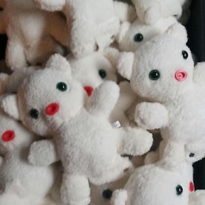 Puede incluir: Un grupo de osos de peluche blancos con ojos verdes y narices rojas. Los osos están dispuestos en una pila, con algunos de ellos parcialmente ocultos por otros.