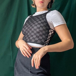 May include: A black crochet top with a lace-up detail. The top is worn over a white tank top and paired with a black skirt.