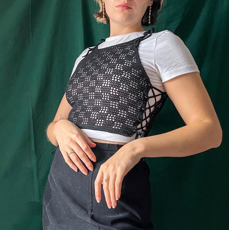 May include: A black crochet top with a lace-up detail. The top is worn over a white tank top and paired with a black skirt.