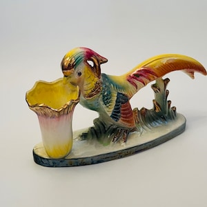 Puede incluir: Figura de faisán de cerámica con un jarrón en forma de flor amarillo, rosa y dorado. El ave tiene un plumaje colorido en tonos azules, amarillos, rojos y verdes. La base ovalada está pintada con hierba.