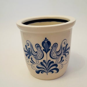Peut inclure: Un pot en céramique crème avec un motif floral et d'oiseaux bleu. Le pot a une forme cylindrique avec un bord légèrement évasé et un intérieur bleu foncé. Le motif présente deux oiseaux face à face, avec des éléments floraux.