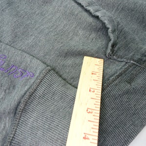 May include: Close-up of a gray sweatshirt with a purple embroidered logo that reads "Southpaw". A wooden ruler is placed next to the sweatshirt to show the size of the cuff.