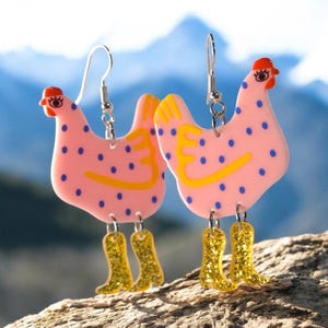 Op de afbeelding: Een paar grillige oorbellen met roze kippenvormen met blauwe stippen en gele accenten. De kippen hebben rode kammen en dragen glitterende gouden cowboylaarzen. De oorbellen hangen aan zilveren haken.