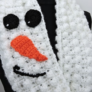 May include: White crocheted infinity scarf with a snowman face. The face has black eyes, an orange nose, and a black smile.