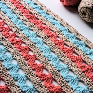 May include: Crocheted blanket in a repeating pattern of coral, light blue, and tan. The yarn is arranged in diagonal stripes. Several balls of yarn in coordinating colors are in the background.